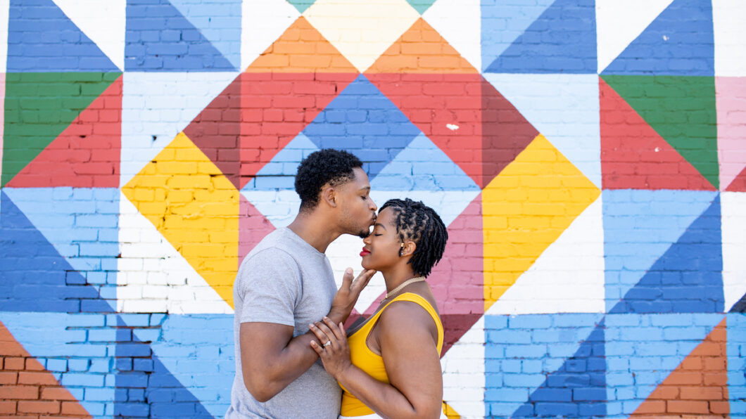 A couple stands in front of a colorful geometric mural. The man kisses the woman's forehead as they hold hands.