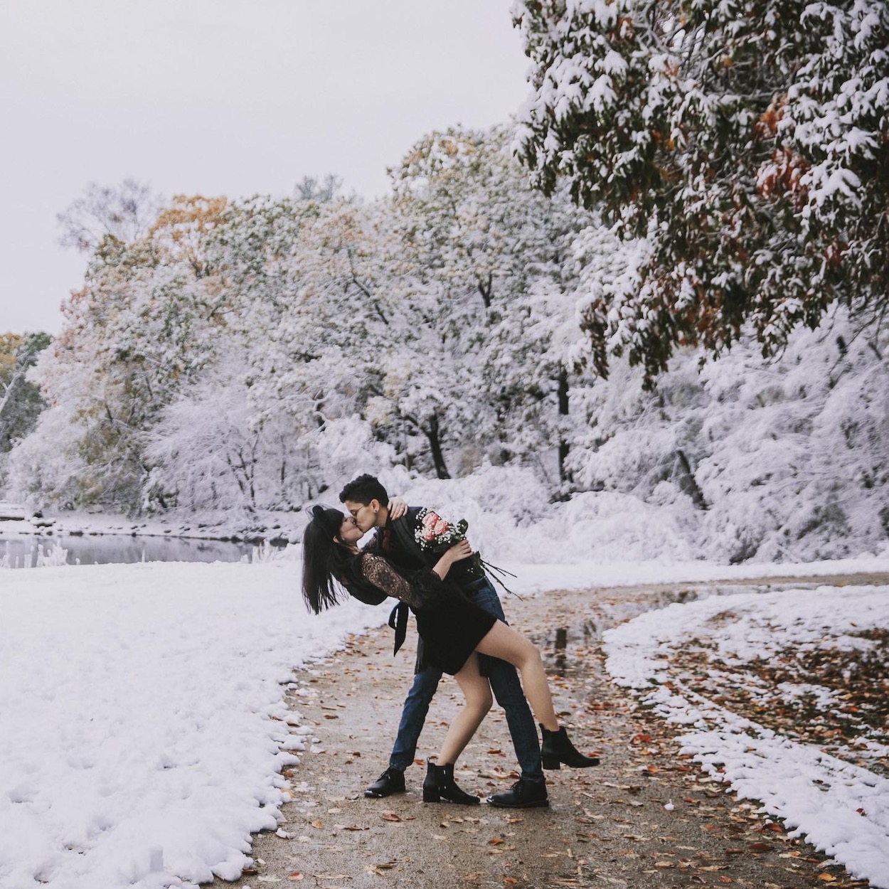 A couple kisses on a snow-covered path lined with trees, with one playfully dipping the other backward.