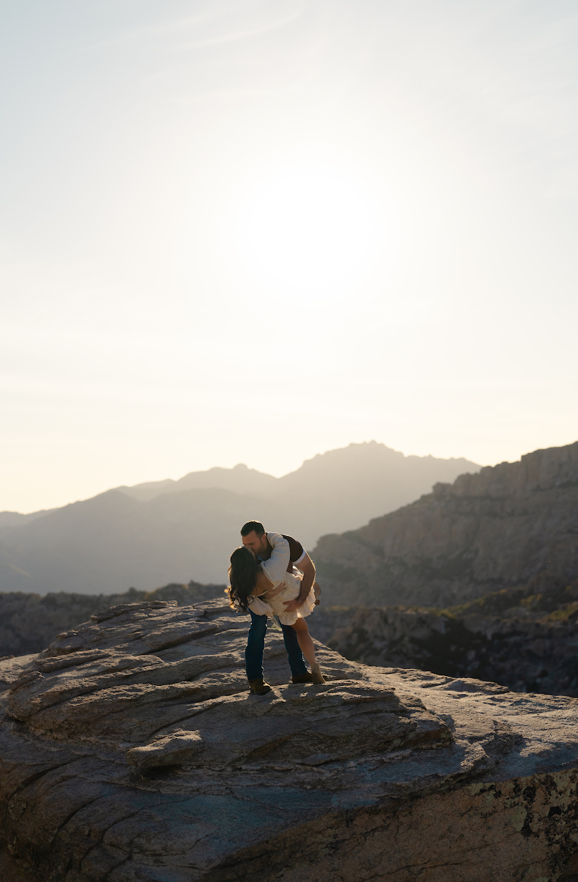 A couple embraces on a rocky ledge with a mountainous landscape and the sun setting in the background.