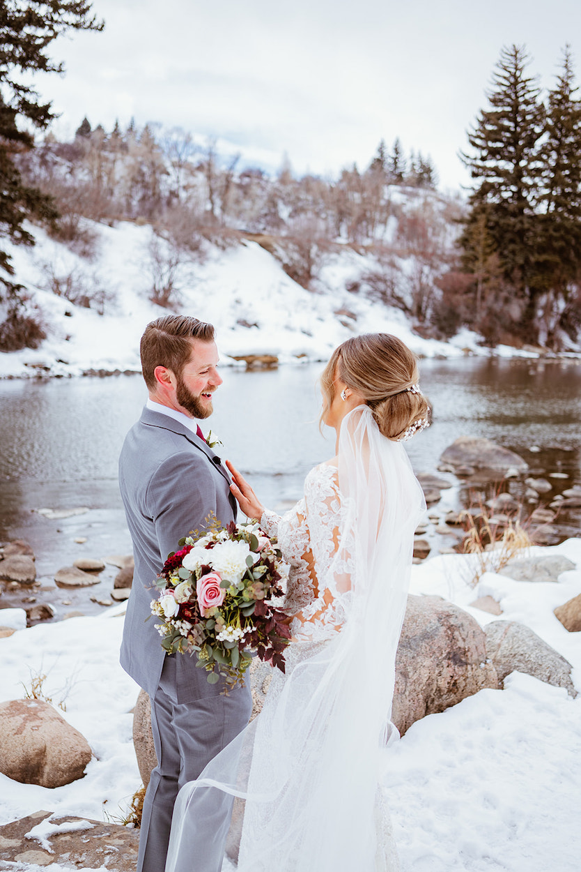 Couple in wedding attire stand by a snowy riverbank. Bride holds a bouquet and wears a veil; groom wears a light gray suit. Snow-covered trees and rocks surround them.