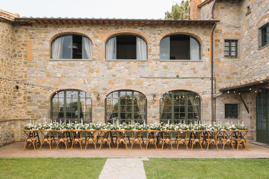 Long dining table set for an outdoor event, with white flowers and candles, placed in front of a rustic stone building.