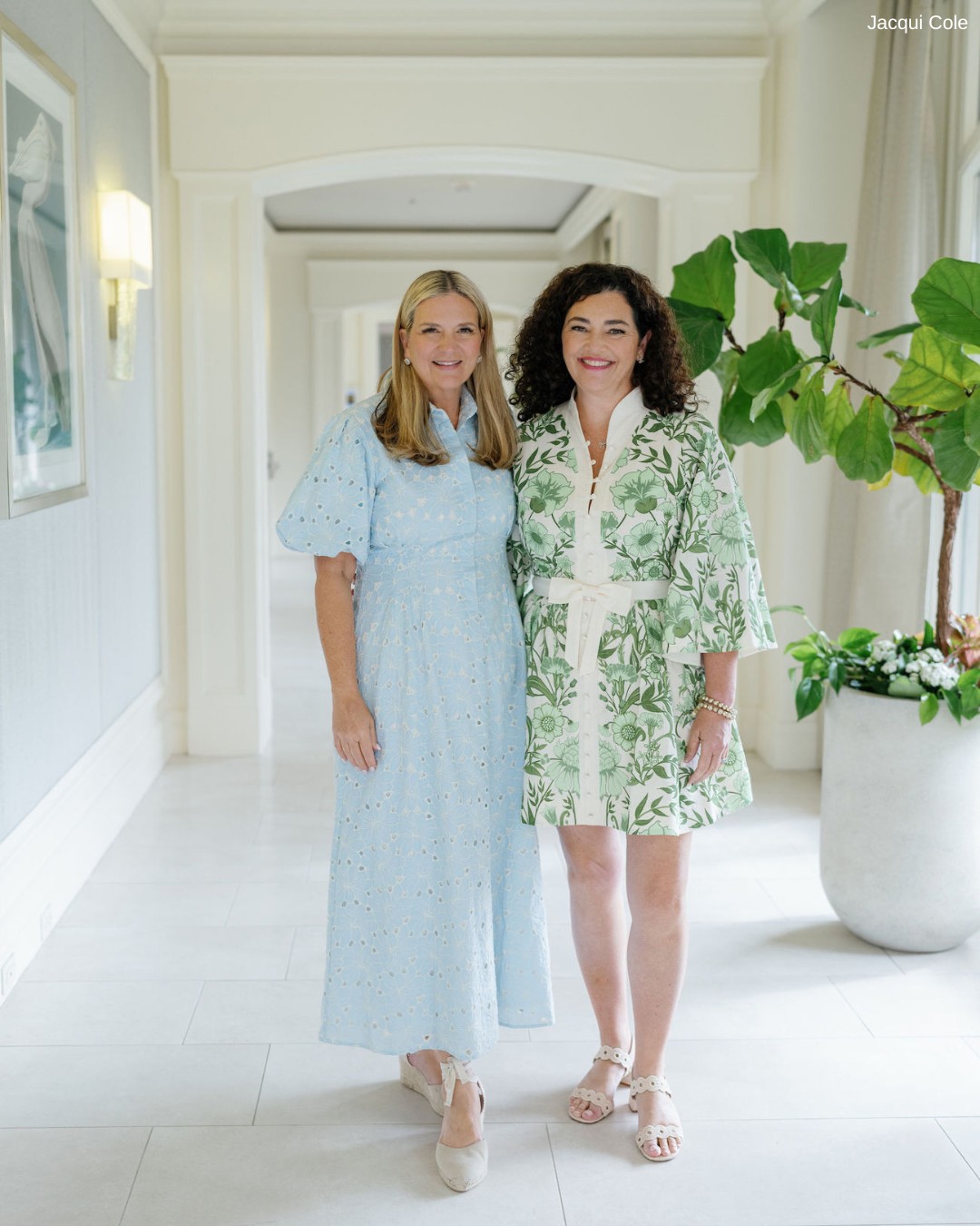 Two women stand together in a bright hallway with pale walls and a large potted plant. One wears a light blue dress; the other wears a green and white floral dress.