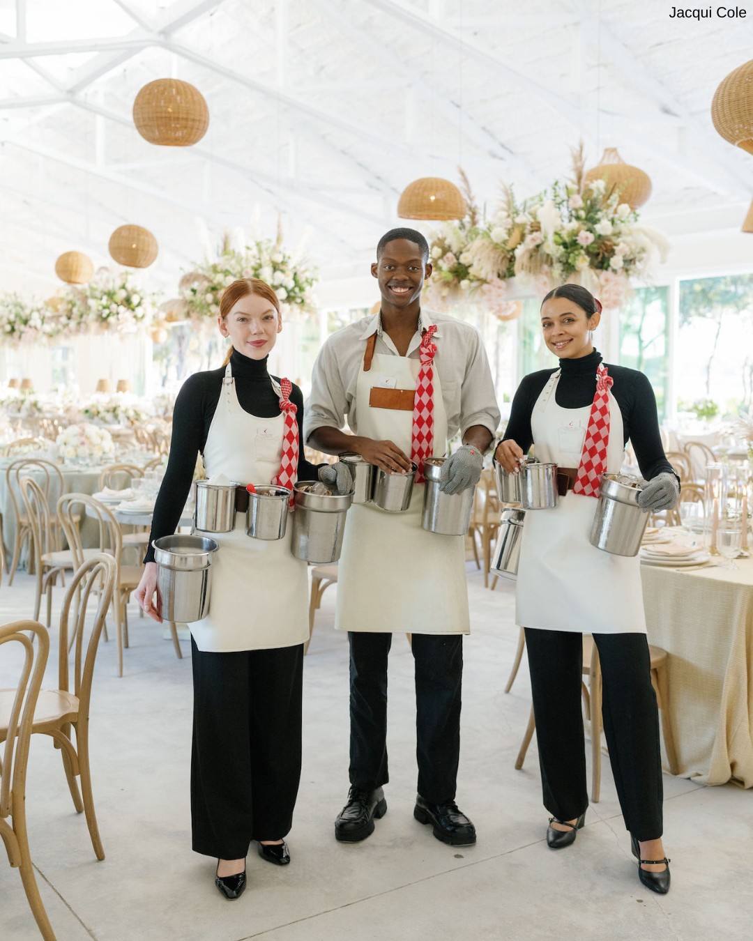 Three people in aprons and checked scarves hold metal canisters of oysters, standing in a decorated venue with tables and floral arrangements.