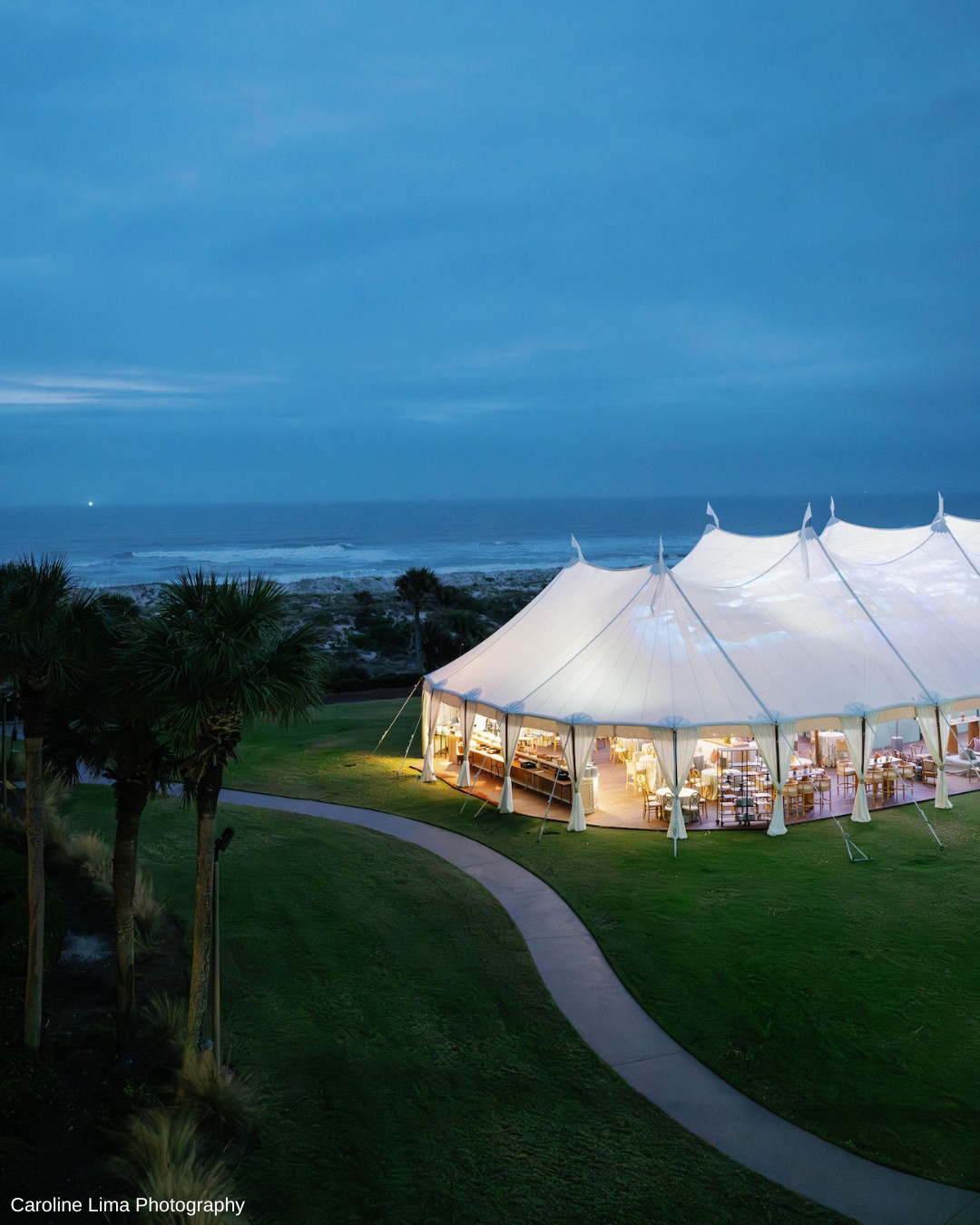 Large white tent illuminated from inside, set on a grassy area near the ocean at dusk, with a winding path and palm trees nearby.