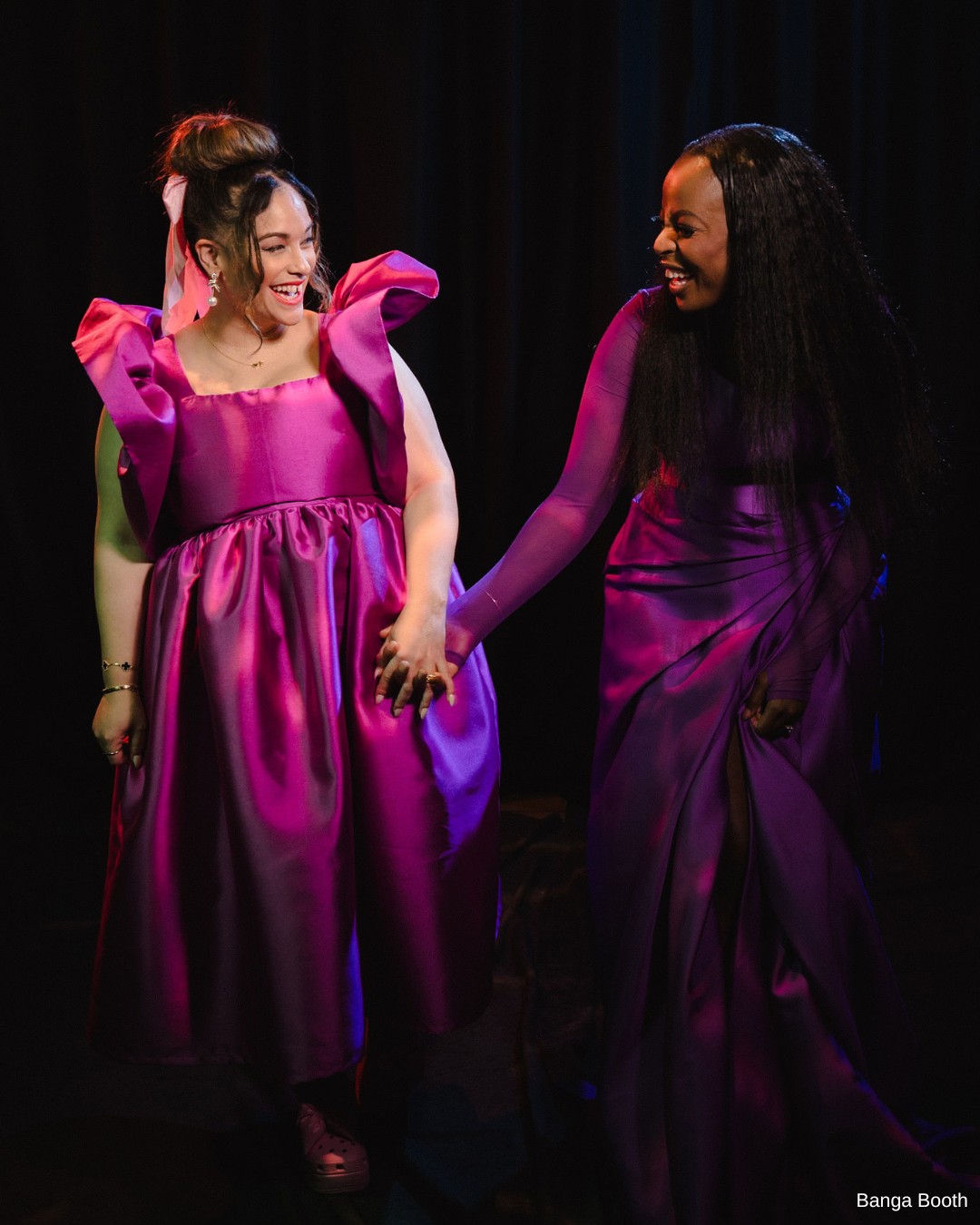 Two people in vibrant purple dresses smile and hold hands against a dark background.