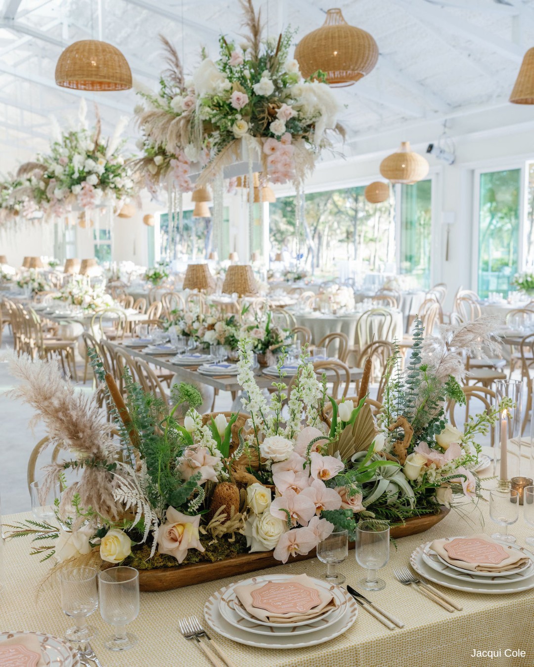 Elegant banquet table with floral centerpiece, cushioned chairs, wicker lighting fixtures, and large windows. Table settings include plates, glasses, and menus on a beige tablecloth.