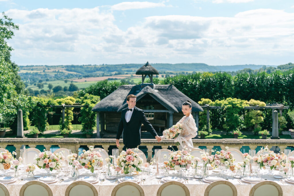 A couple in formal attire poses in a manicured garden with a pool and wooden pavilion.