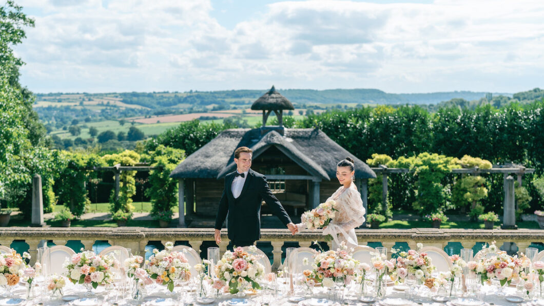 A couple in formal attire poses in a manicured garden with a pool and wooden pavilion.