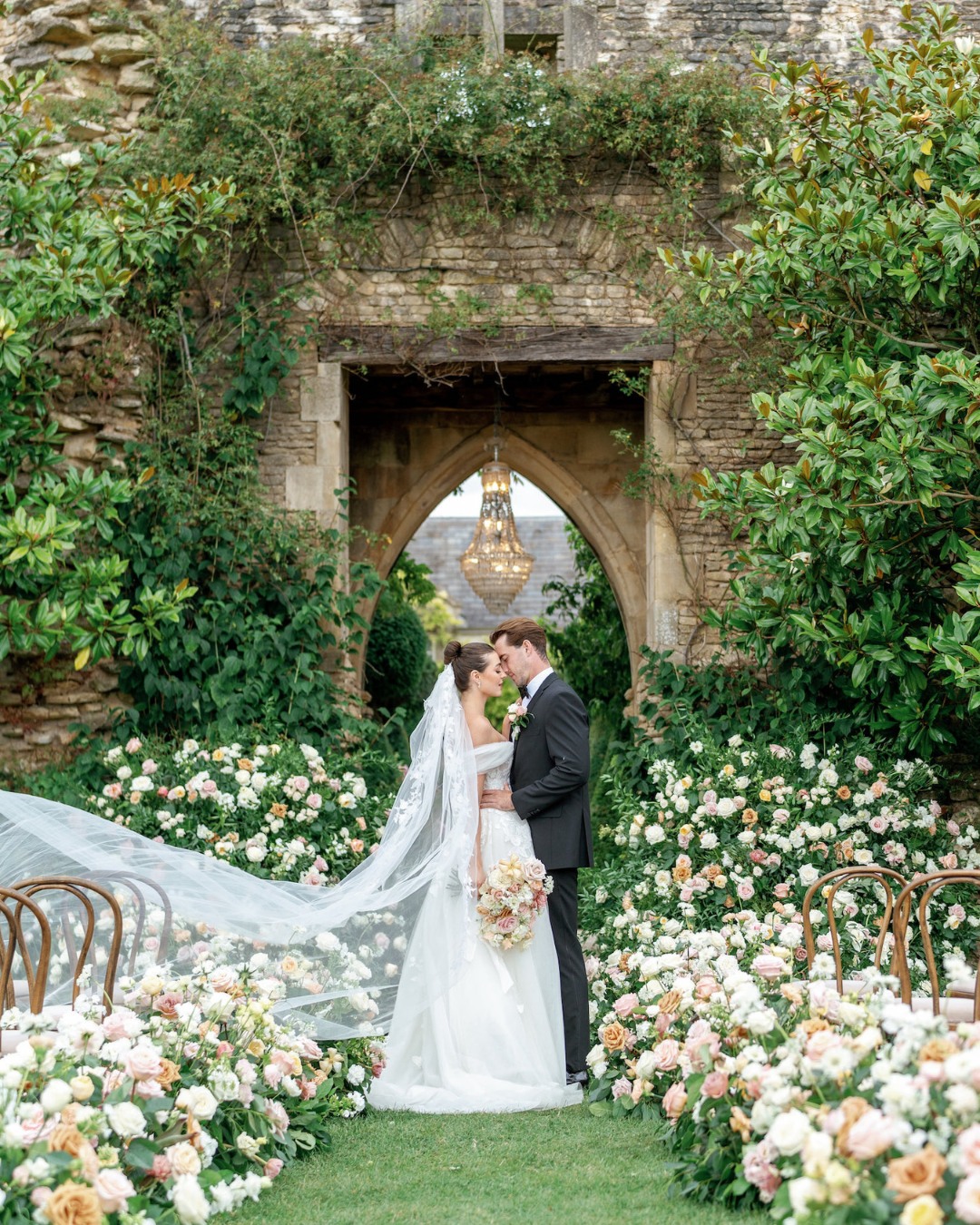 Bride and groom stand in a lush garden surrounded by flowers, beneath an archway with a hanging chandelier. The bride wears a flowing white gown with a veil, and the groom is in a black suit.