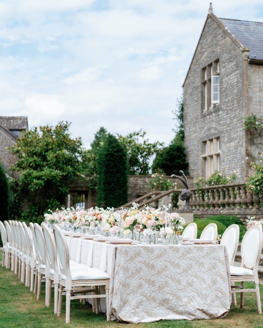 Outdoor table setting with elegant white and floral decor in a garden, next to a stone building under a cloudy sky.