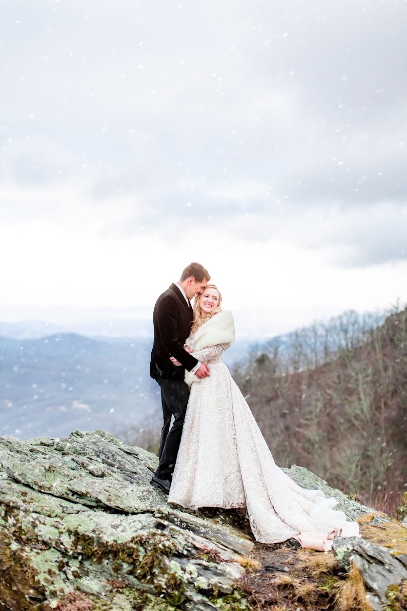 A bride and groom stand on a rocky hilltop in snowfall. The bride wears a long gown and shawl, while the groom is dressed in a dark suit. The background features a mountainous landscape.