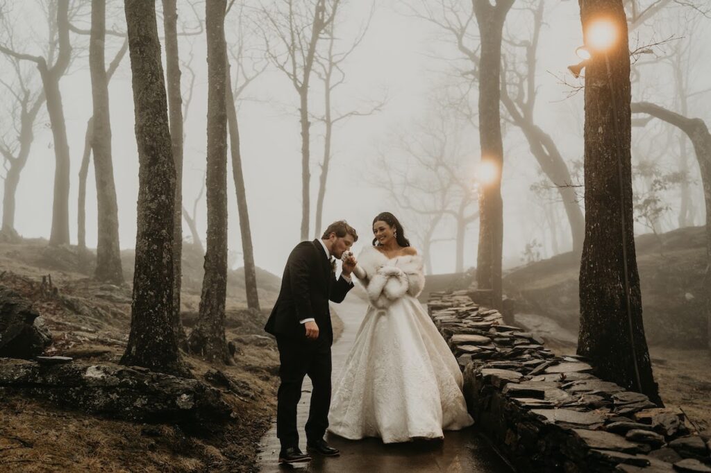 A couple in formal attire walks in a misty forest. The woman wears a white gown and fur stole, while the man dons a suit. They walk along a stone path surrounded by leafless trees.