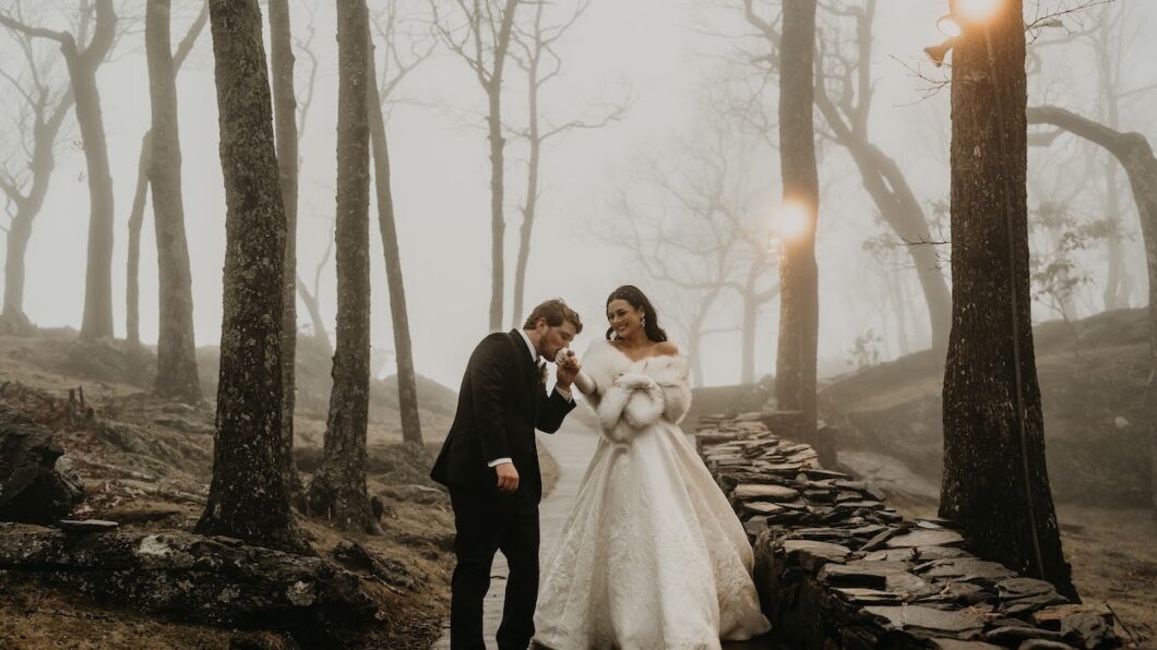 A couple in formal attire walks in a misty forest. The woman wears a white gown and fur stole, while the man dons a suit. They walk along a stone path surrounded by leafless trees.