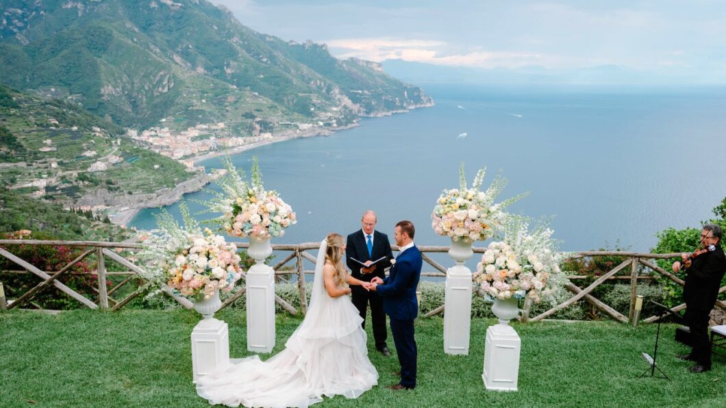 Rosaleny & Cameron - Ravello, Italy - Featured- Website (1350 x 1080 px) (1) Outdoor wedding ceremony overlooking a coastal landscape. A couple stands before an officiant, surrounded by flower arrangements.