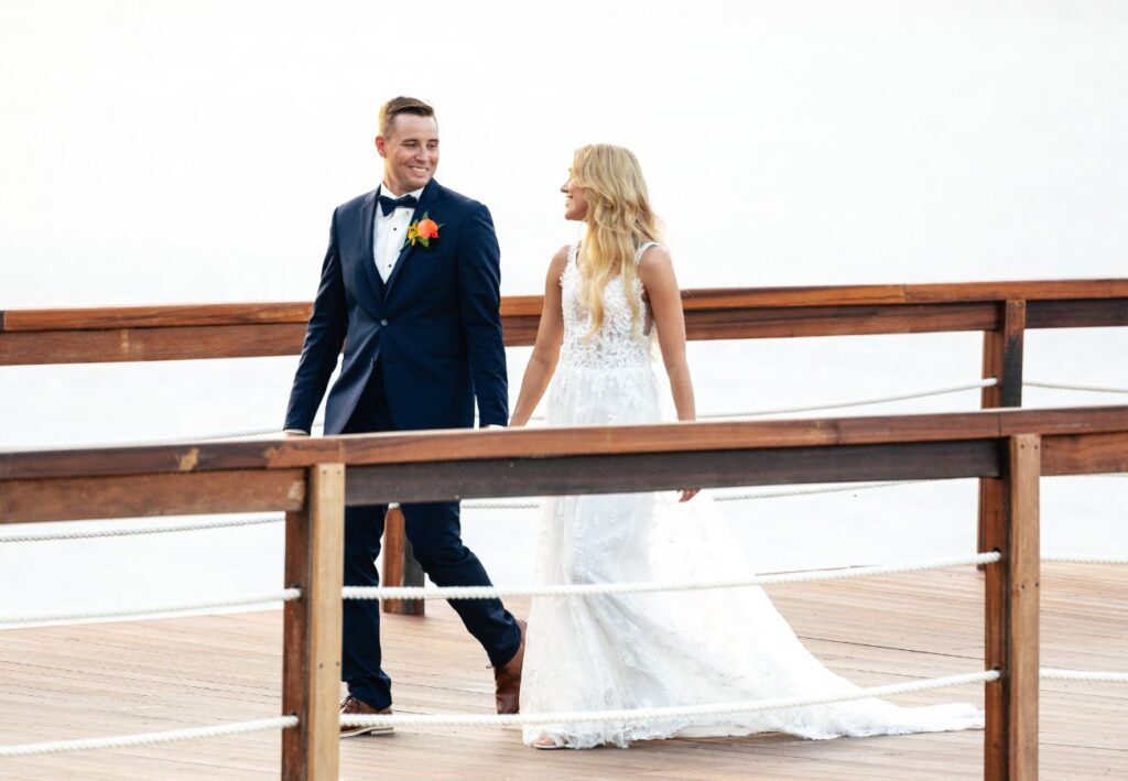 A couple in wedding attire walks hand in hand on a wooden deck.