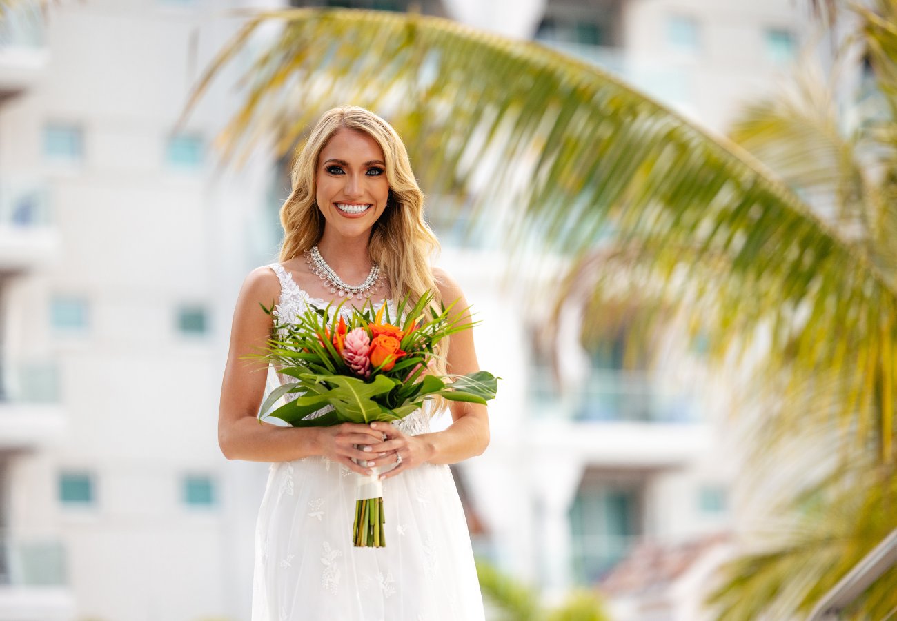 A woman in a white dress holds a bouquet of flowers, standing outside near palm fronds with a building in the background.