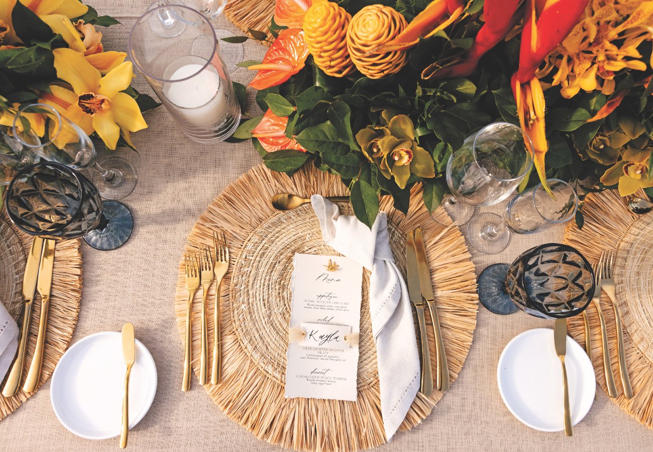 Elegant table setting with tropical flowers, round woven placemats, black glassware, gold cutlery, and small menus on each plate.