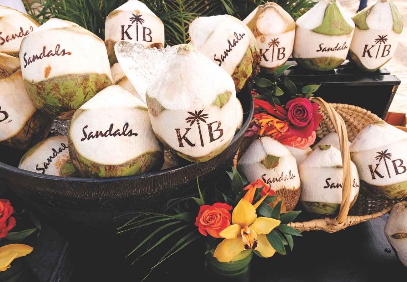 Several coconuts branded with "KB" and "Sandals" are displayed in a basket and on a table, surrounded by colorful flowers.