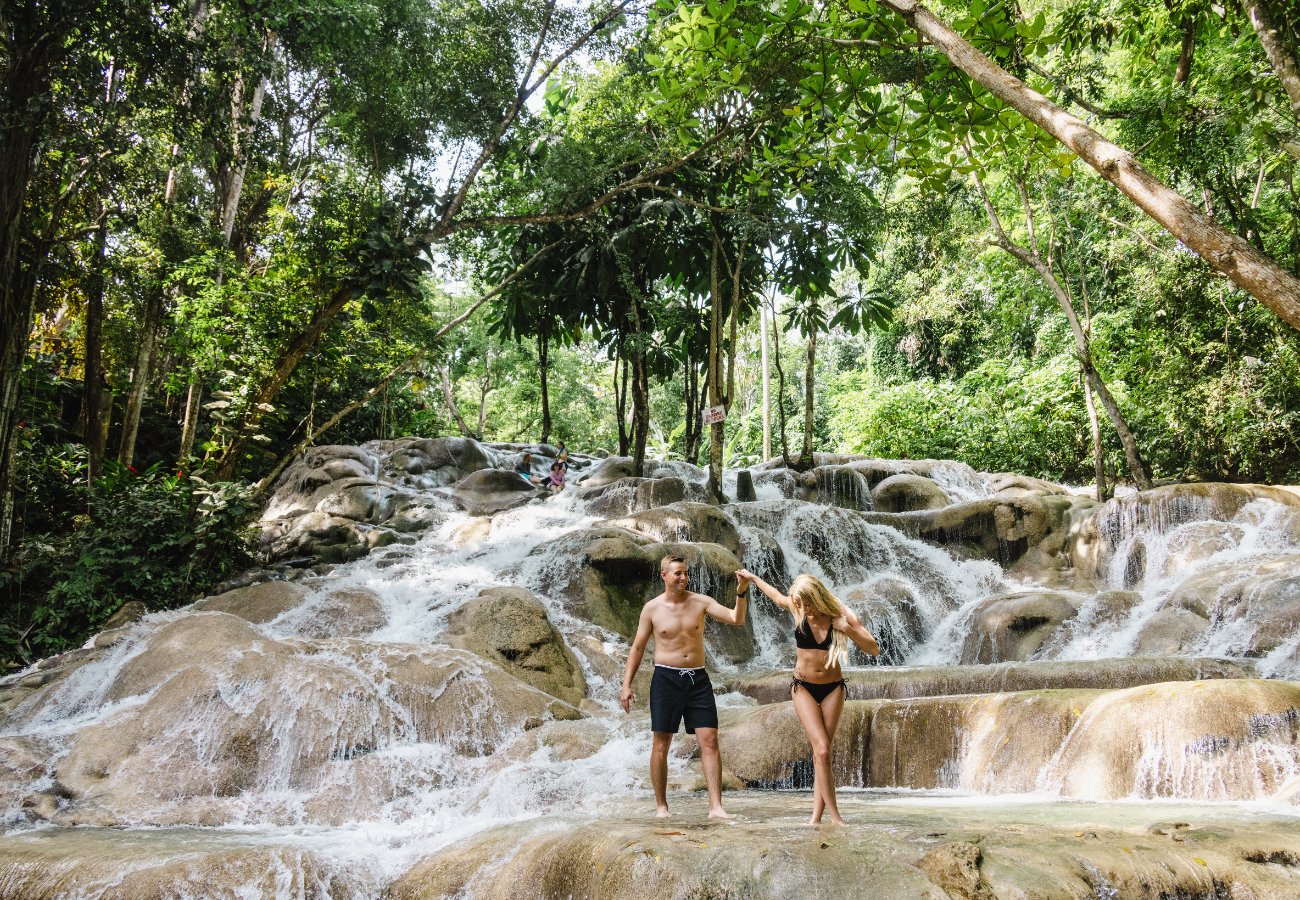 A man and woman in swimwear hold hands while walking on rocky waterfalls surrounded by lush green trees.