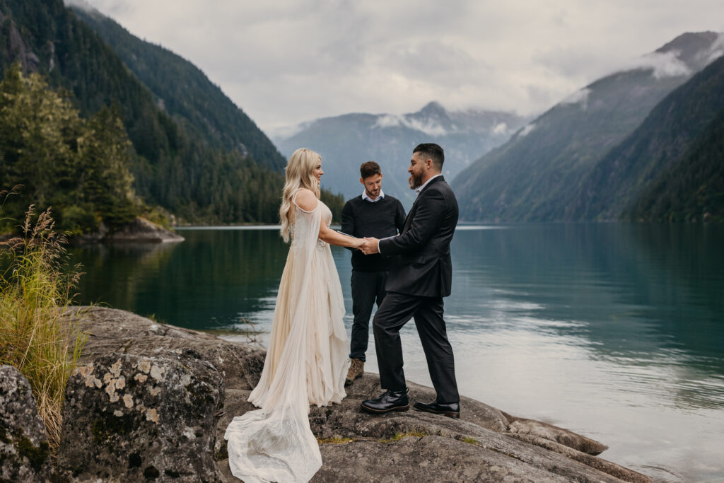 A couple is getting married outdoors by a lake, surrounded by mountains, while an officiant conducts the ceremony.