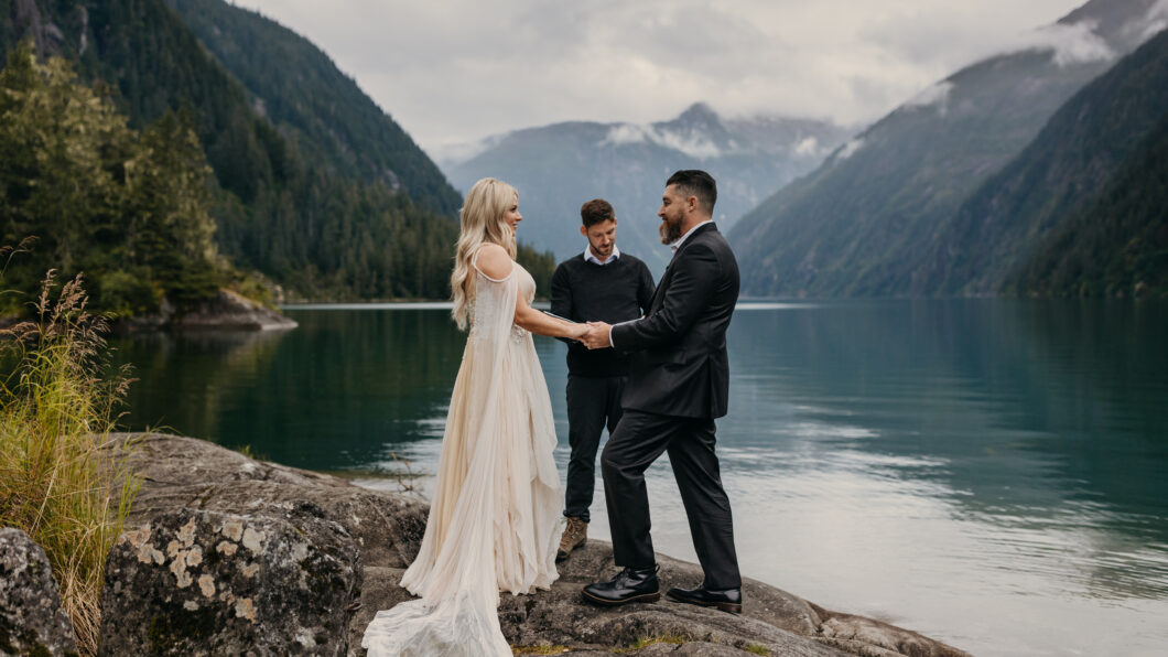 A couple is getting married outdoors by a lake, surrounded by mountains, while an officiant conducts the ceremony.