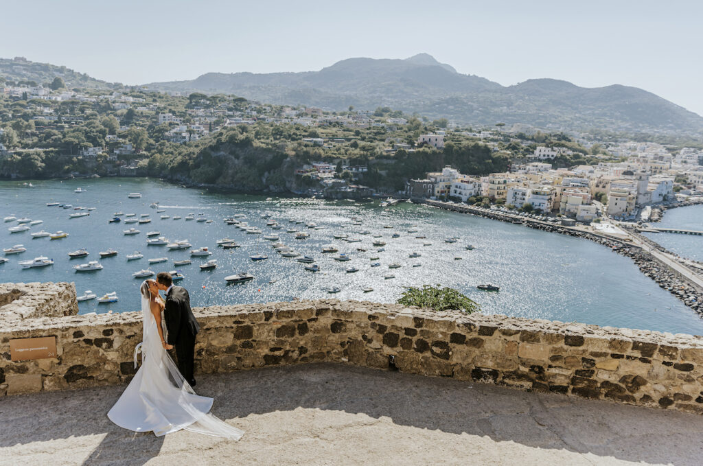 A bride and groom embrace on a stone terrace overlooking a coastal village and bay filled with boats on a sunny day.