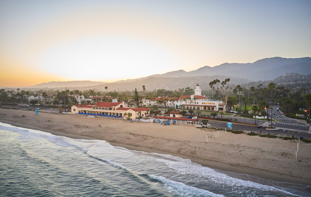 Aerial view of a beach at sunset with a coastal town, palm trees, and mountains in the background.