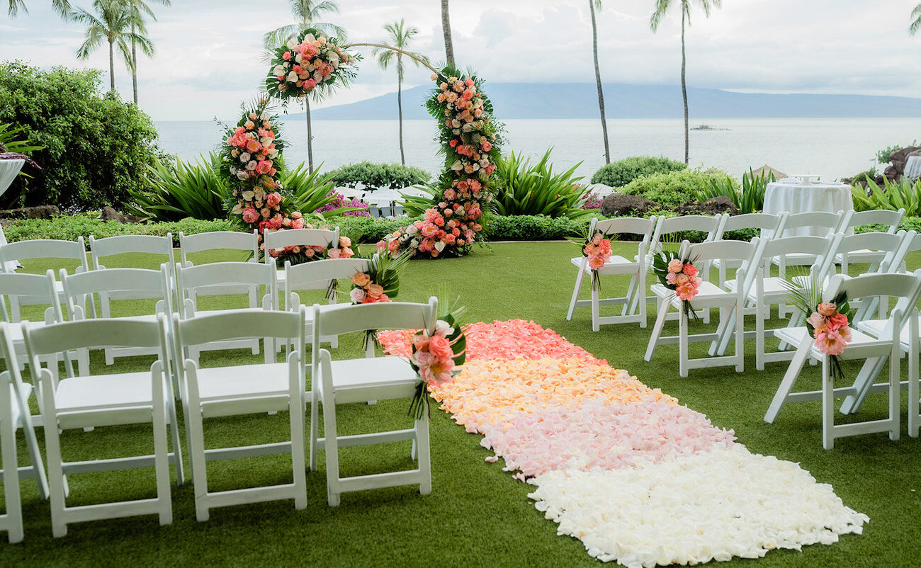 Outdoor wedding setup with white chairs, a circular floral arch, and a petal-covered aisle leading towards the sea, set against a backdrop of palm trees and mountains.