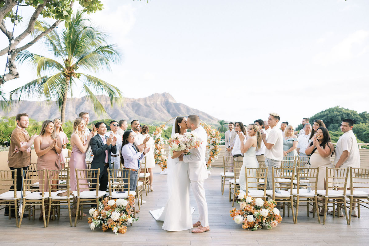 A couple kisses at an outdoor wedding ceremony with guests applauding. There are floral arrangements and mountains in the background.