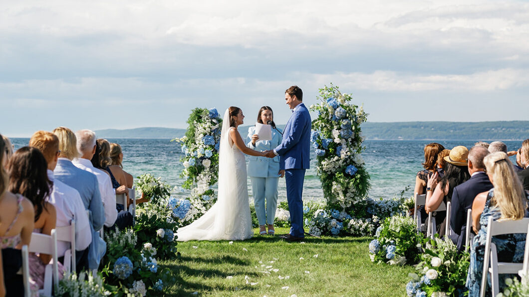 A bride and groom stand facing each other at an outdoor wedding ceremony by the sea. A person officiates, and guests are seated nearby. Blue floral decorations surround the couple.