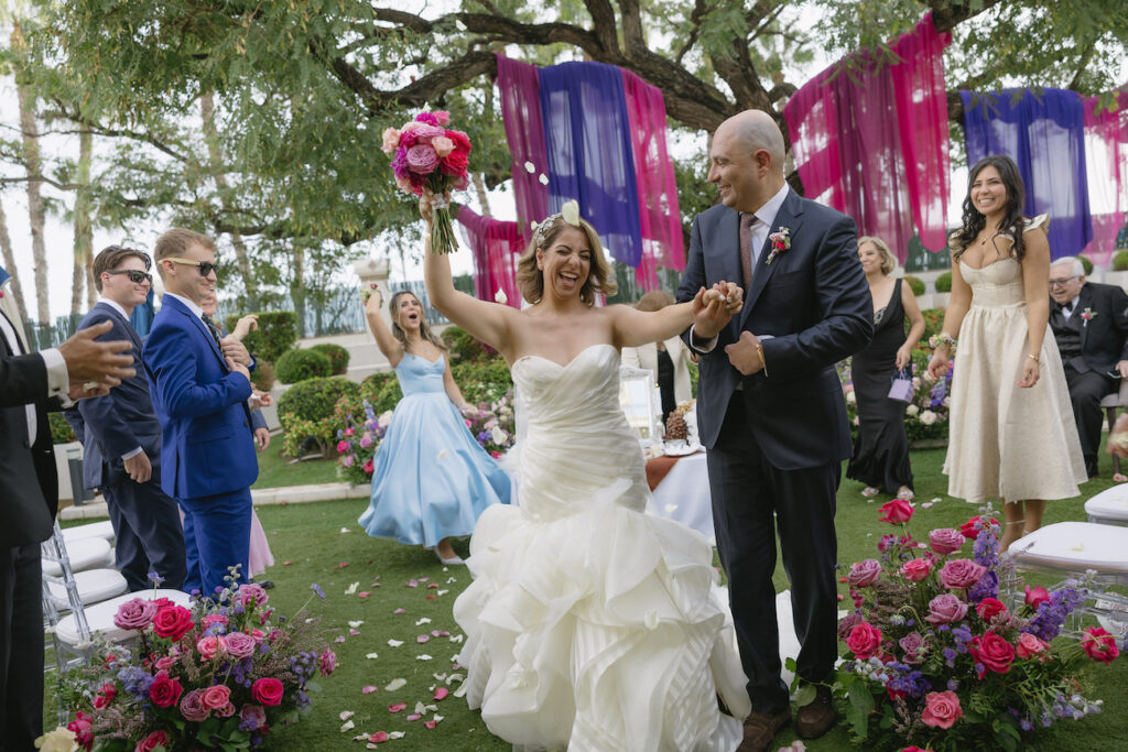 Bride and groom walk joyfully down the aisle outdoors, showered with petals. Guests cheer them on, with vibrant purple and pink decorations in the background.