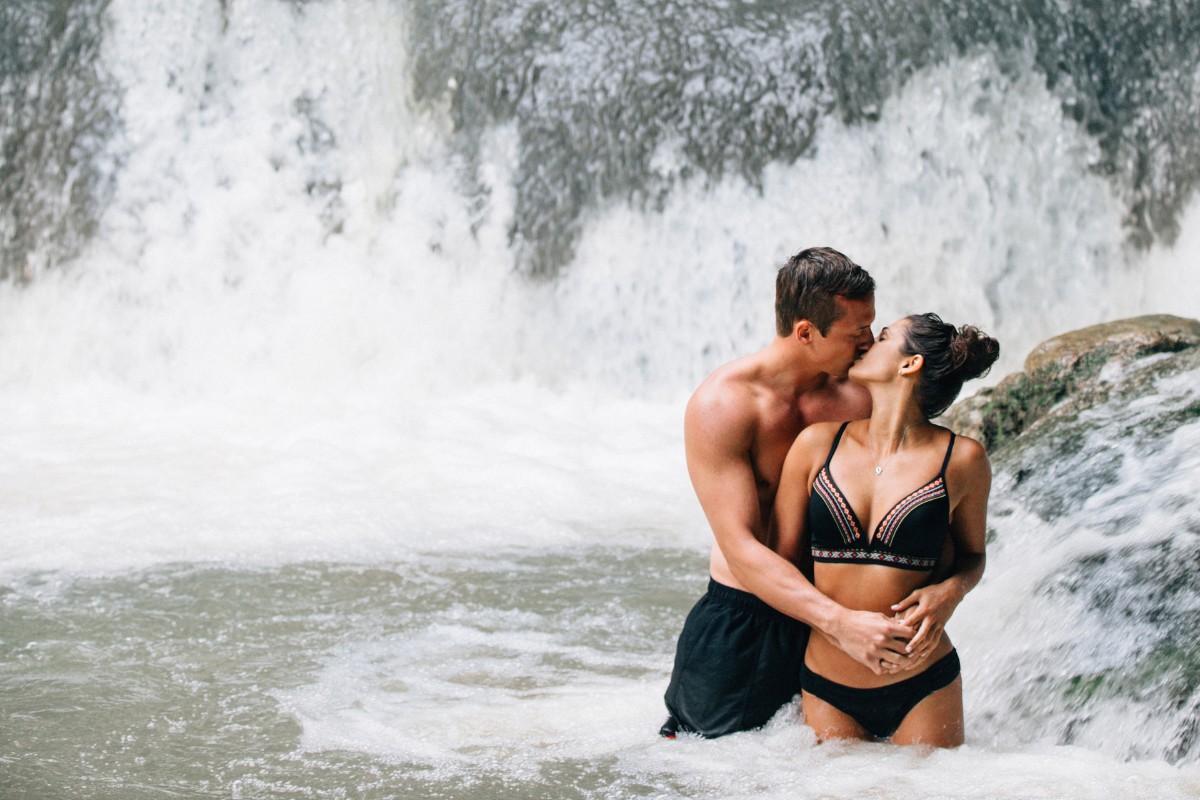 A couple in swimwear kisses while standing in a river with a waterfall in the background.