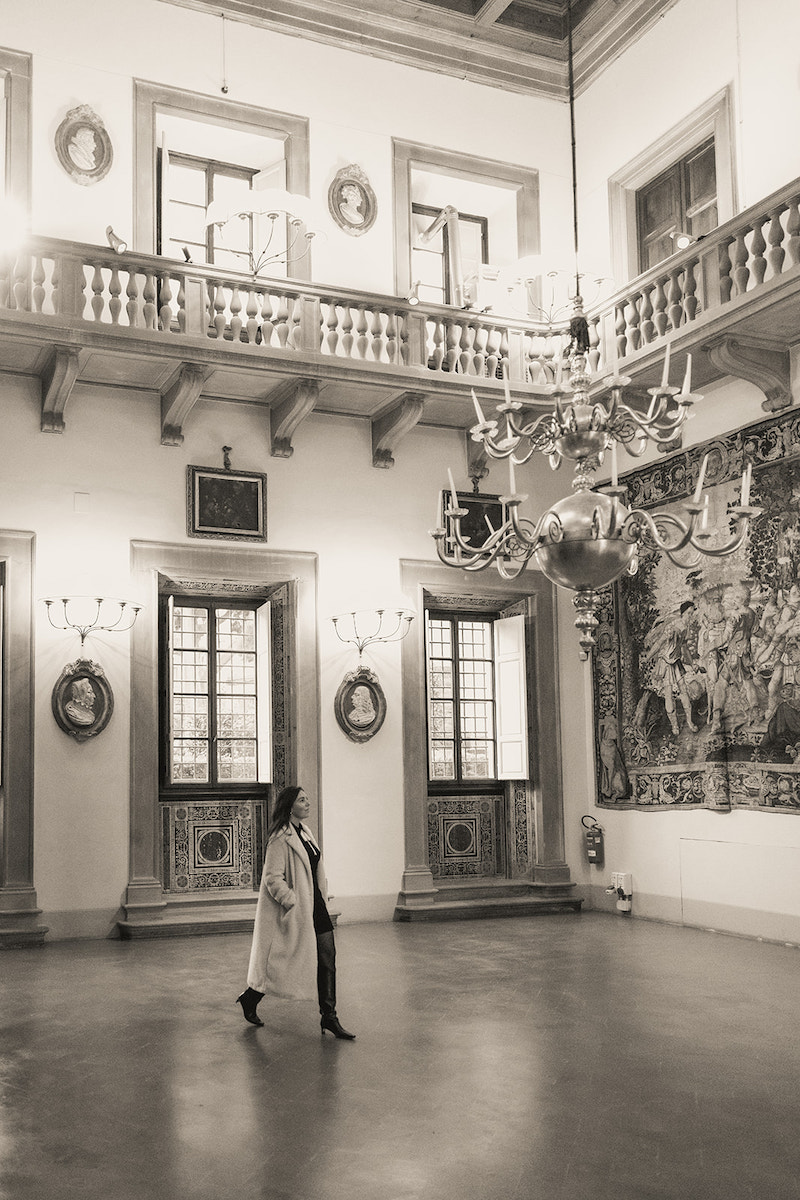 A person in a long coat walks across an ornate, high-ceilinged room with large windows, chandeliers, and a tapestry on the wall.