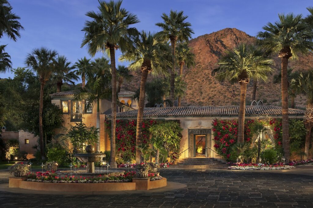 A Mediterranean-style villa with palm trees, flowering vines, and a courtyard fountain sits at the base of a rocky mountain at dusk.