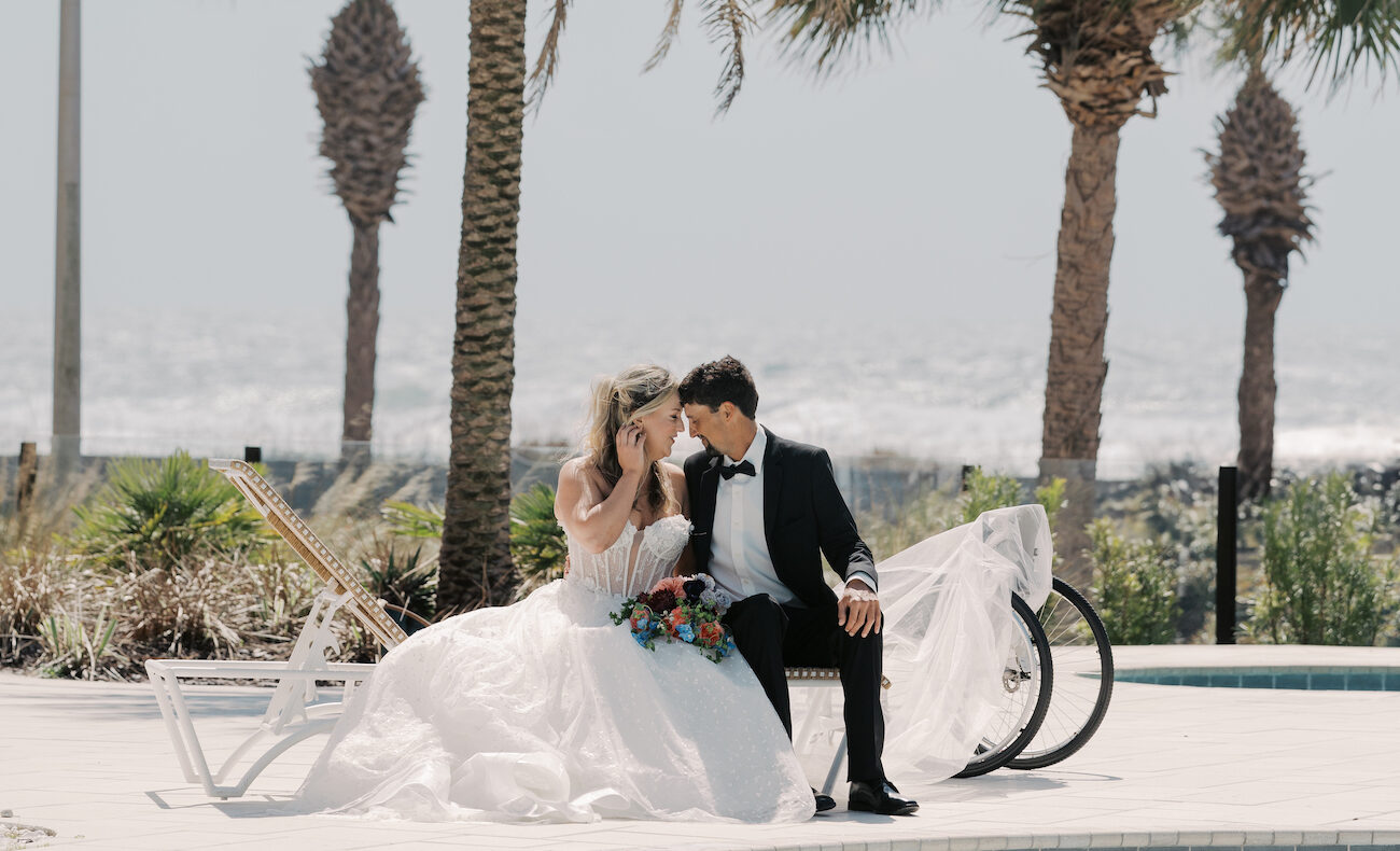 A bride and groom sit together outdoors near palm trees and a bicycle, with the ocean in the background.