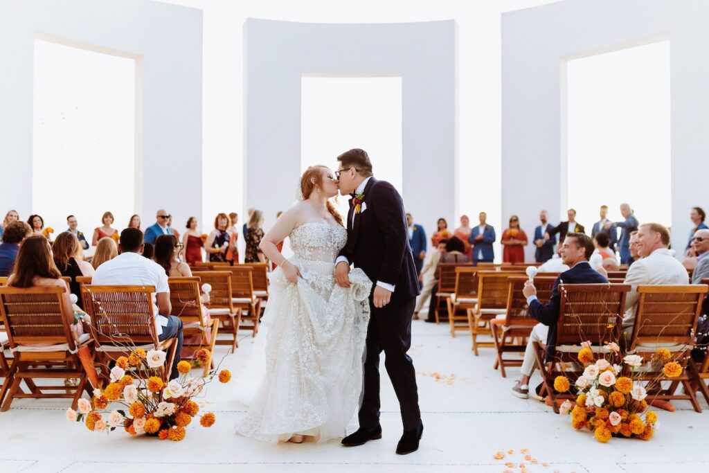 A bride and groom kiss at an outdoor wedding ceremony, surrounded by guests seated on wooden chairs with orange and yellow floral decorations.