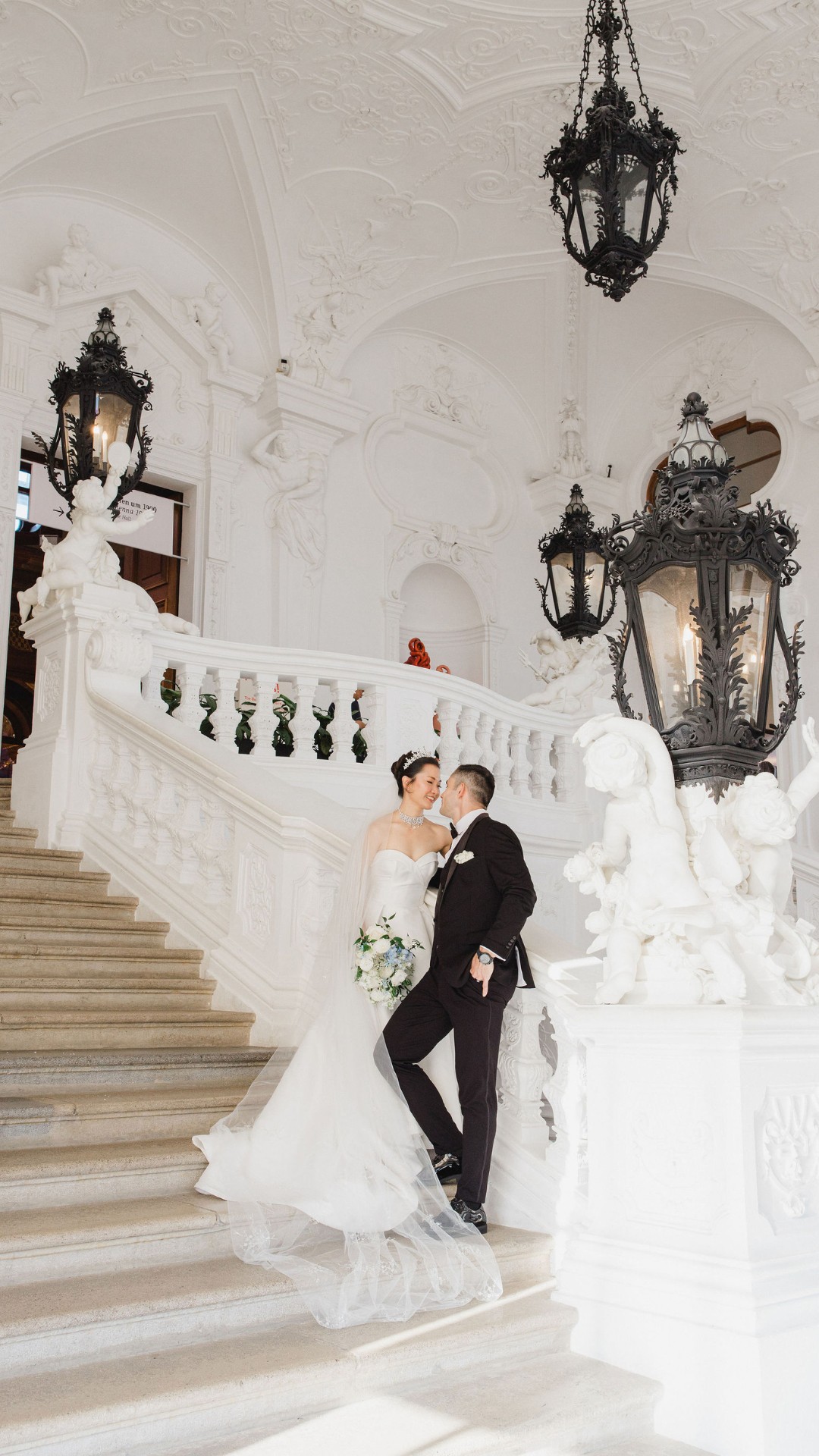 A bride and groom stand on an ornate staircase in a grand, white interior with elaborate decorations and large black lanterns.