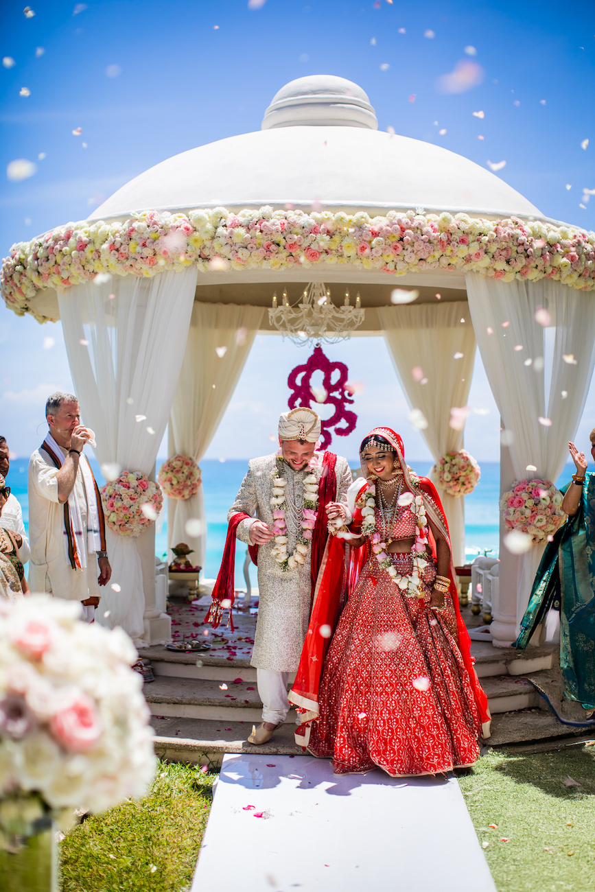 A bride and groom in traditional Indian attire walk under a flower-adorned wedding mandap by the seaside, surrounded by guests.