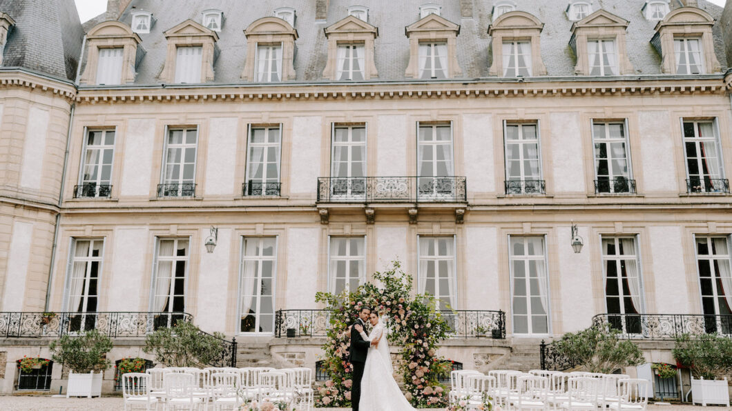 A bride and groom stand together in front of a floral arch, outside a grand building with empty chairs arranged for a wedding ceremony.