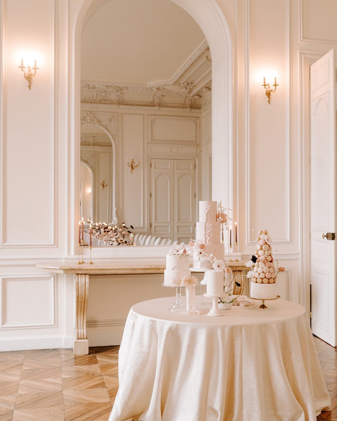 A round table with elegant white cakes and pastries stands in a softly lit, ornate room with cream-colored walls, large mirrors, and gold sconces.