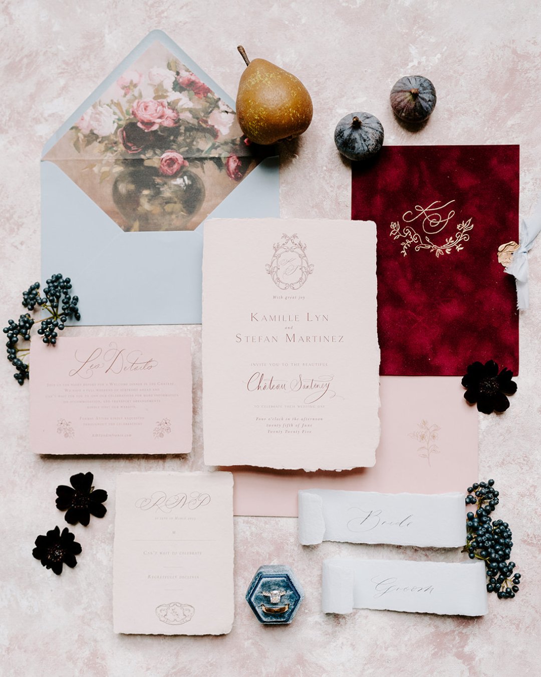 Flat lay of elegant wedding invitations, RSVP cards, envelopes, and name cards arranged with figs, grapes, a pear, and black flowers on a textured light pink background.