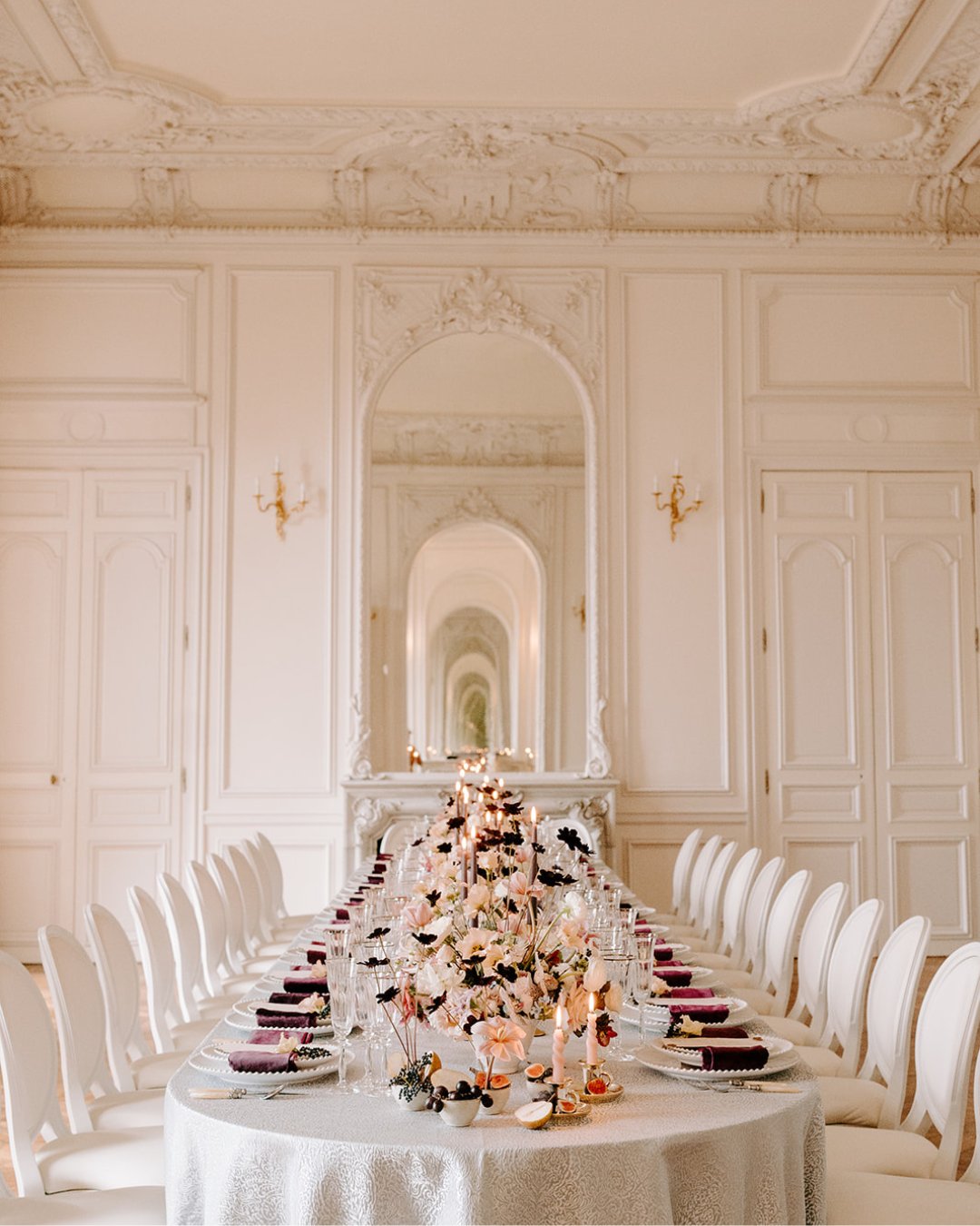 Elegant dining table set for an event in a lavish, ornate room with white walls, decorative molding, and a large mirror reflecting the scene.