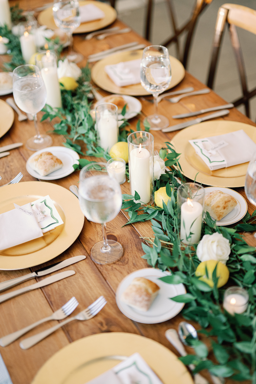 A wooden table set for a meal with yellow plates, bread rolls, water glasses, white napkins, candles, green leaves, and lemons as decorations.