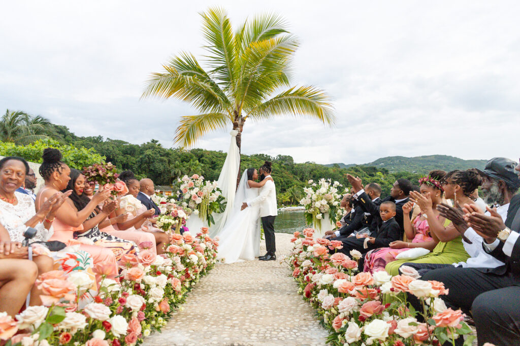 A couple kisses at an outdoor wedding ceremony, surrounded by guests seated along an aisle lined with flowers, with a palm tree and greenery in the background.