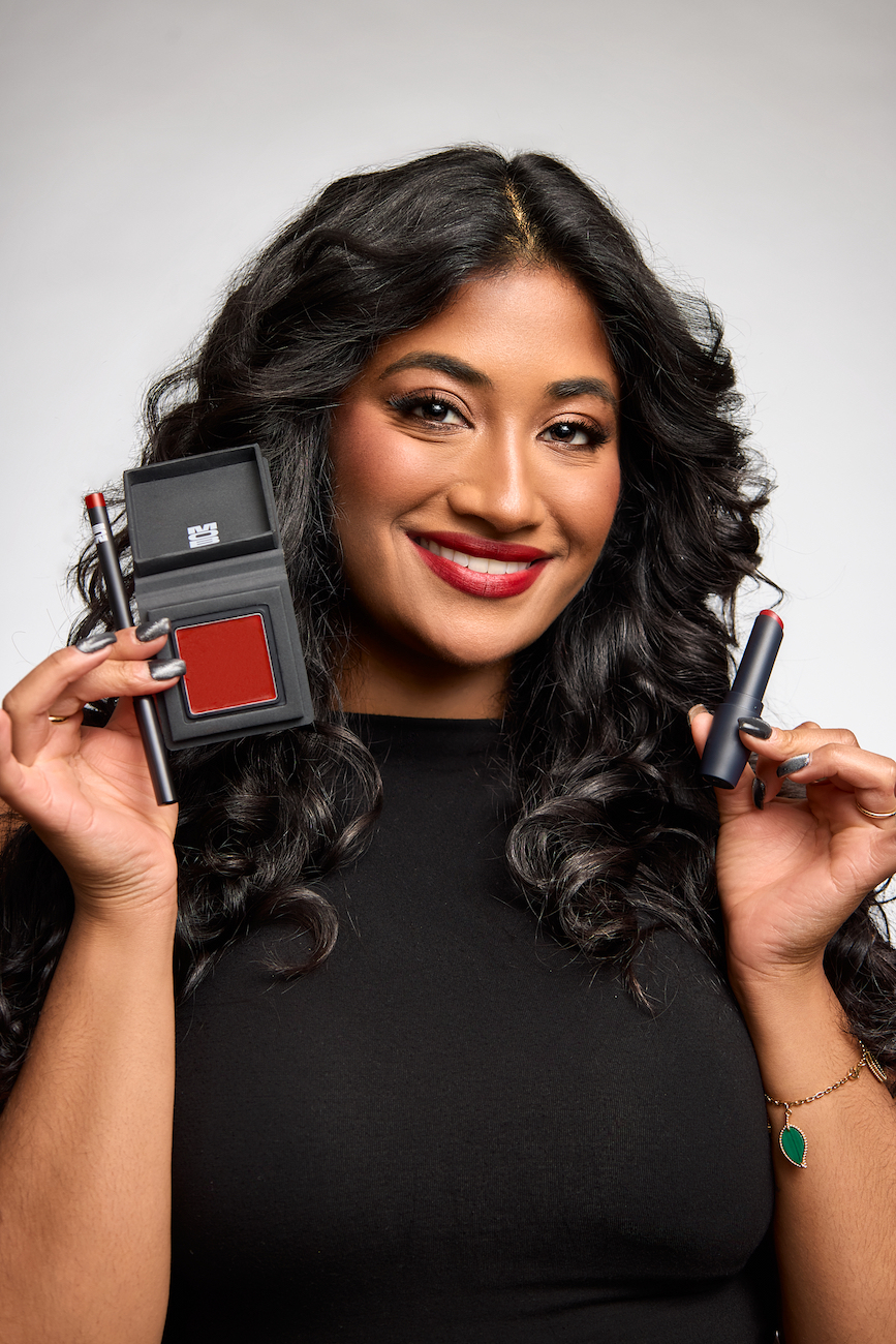 Woman with long dark hair smiles while holding a makeup palette, pencil, and lipstick; she is wearing a black top and red lipstick.