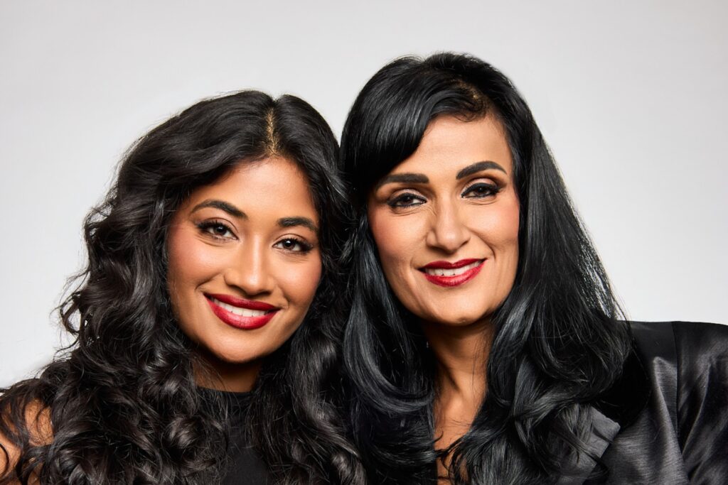 Two women with long black hair and red lipstick smile, standing close together in front of a plain light background.