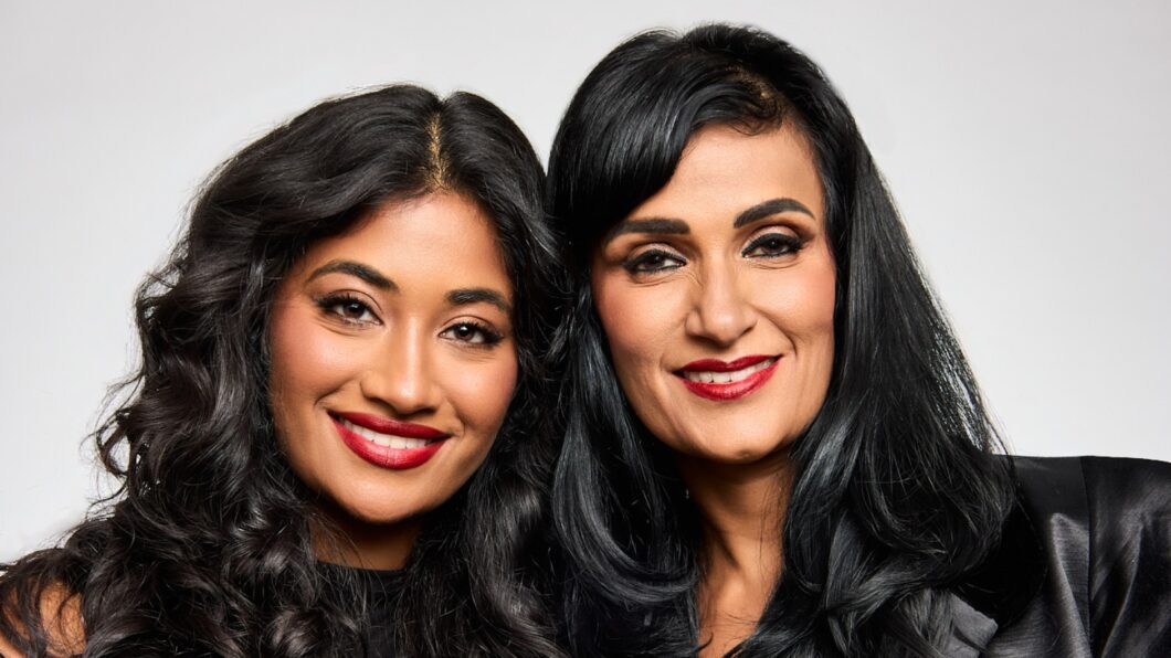 Two women with long black hair and red lipstick smile, standing close together in front of a plain light background.
