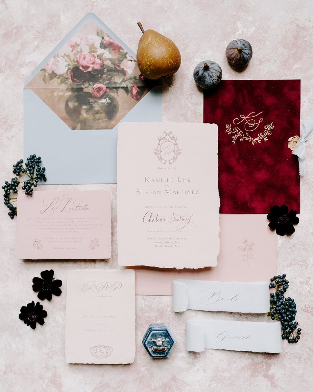 A flat lay of elegant wedding stationery with floral designs, a red envelope, a pear, figs, dark flowers, a blue ring box, and two ribbon name tags on a textured surface.