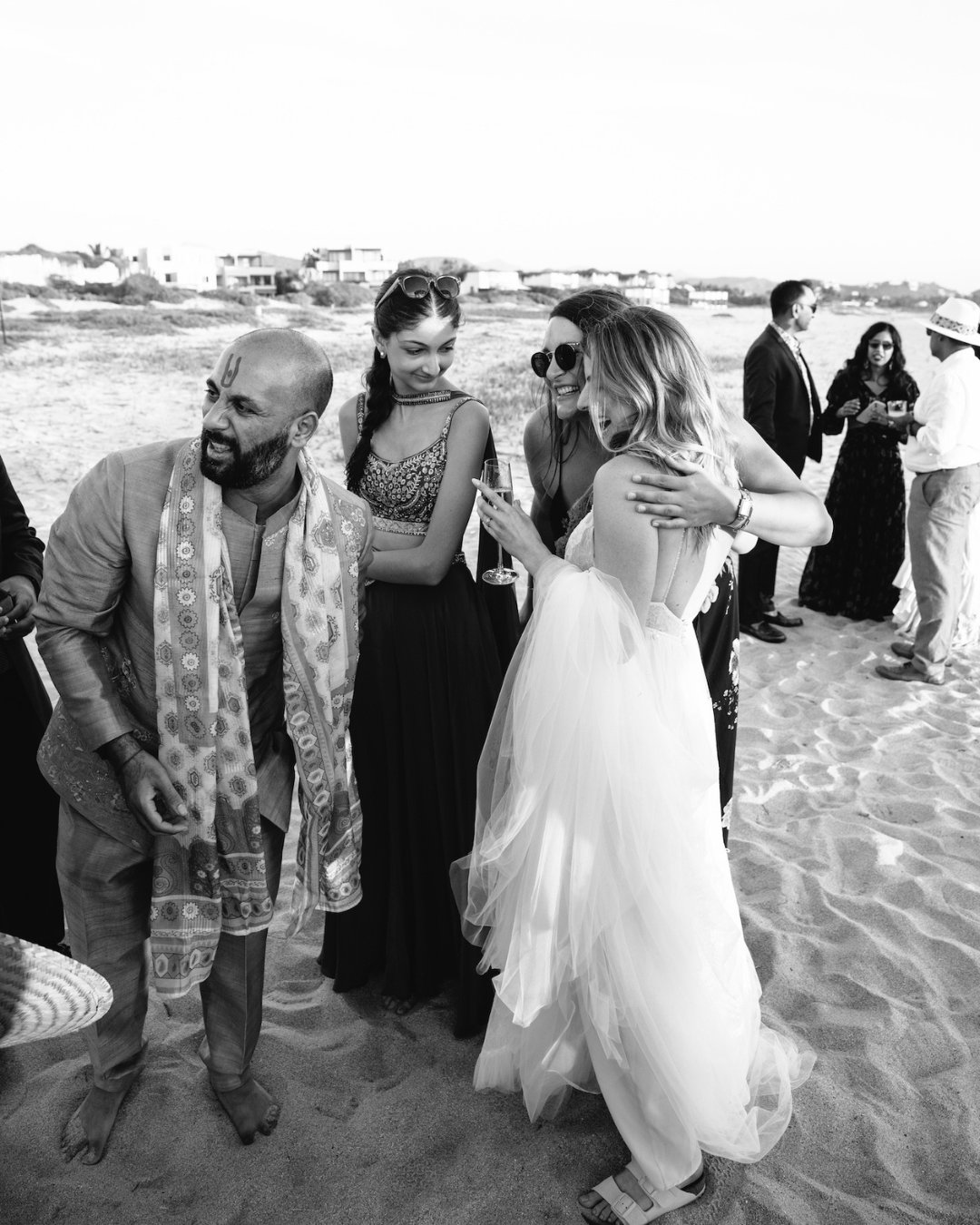 A group of people dressed in formal attire stand and interact on a sandy beach during a social event.