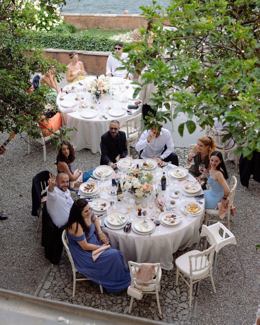 A group of people sit around two round tables set with food and drinks in an outdoor garden setting, viewed from above.